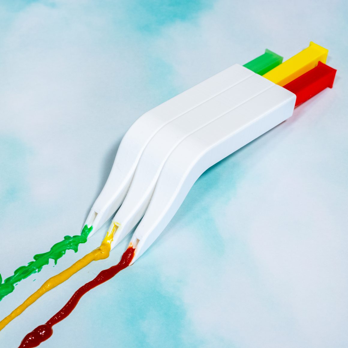 A white fork-shaped gadget dispensing green, yellow and red condiments from separate channels.