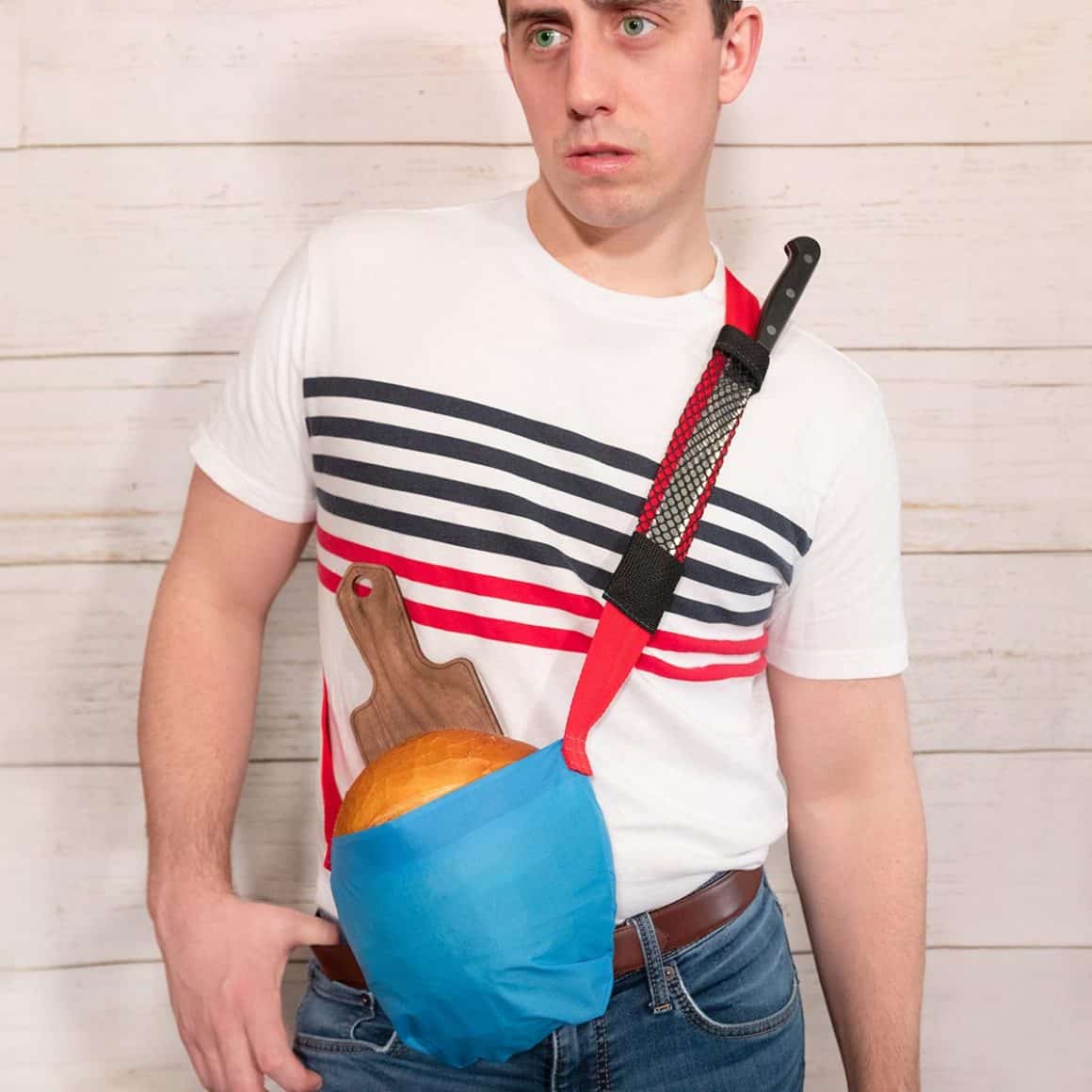 Matty wearing a blue shoulder pouch holding bread with a cutting board and knife alongside.