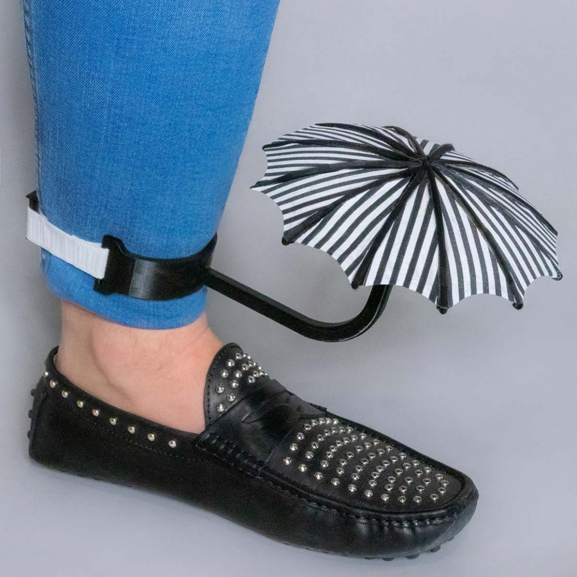 A striped miniature umbrella mounted above a black shoe with a strap around the ankle.